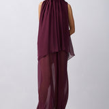 A model wearing a sleeveless, high-neck, draped maxi dress in a deep plum purple color, standing against a light gray studio background.