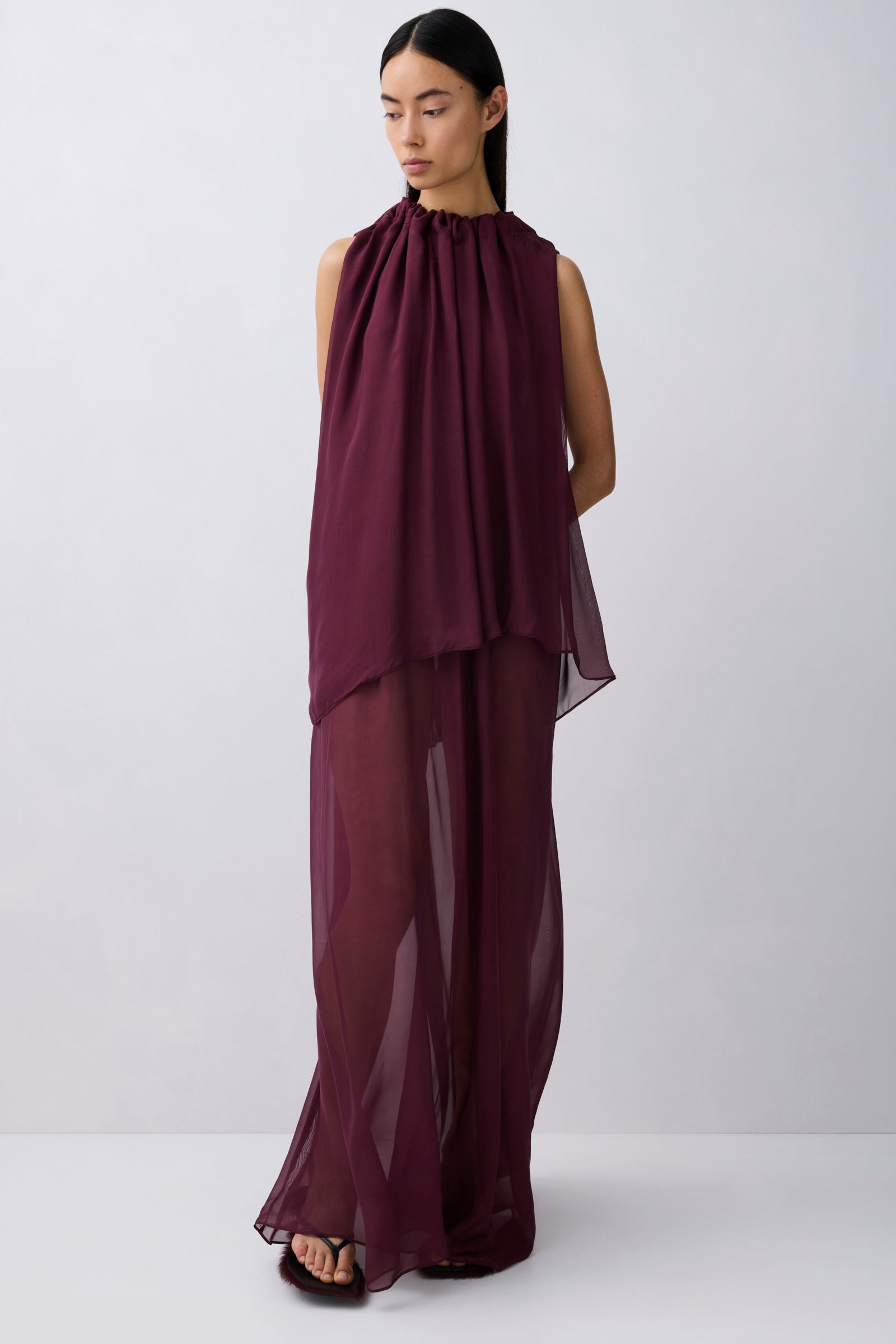 A model wearing a sleeveless, high-neck, draped maxi dress in a deep plum purple color, standing against a light gray studio background.