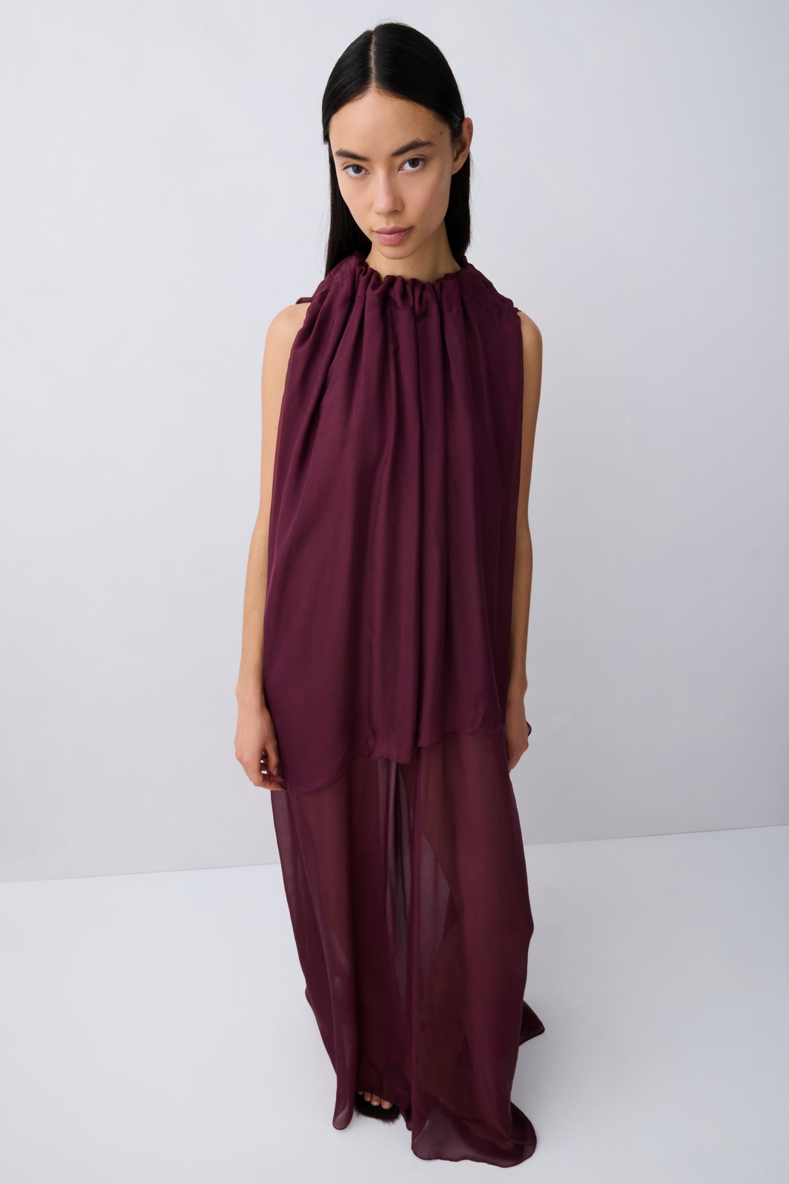 A model wearing a sleeveless, high-neck, deep burgundy maxi dress with a sheer, flowing overlay, photographed against a neutral light gray studio background.