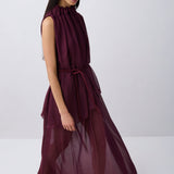 A woman modeling a sleeveless, high-neck, deep burgundy maxi dress with a matching waist tie, set against a plain light gray studio background.