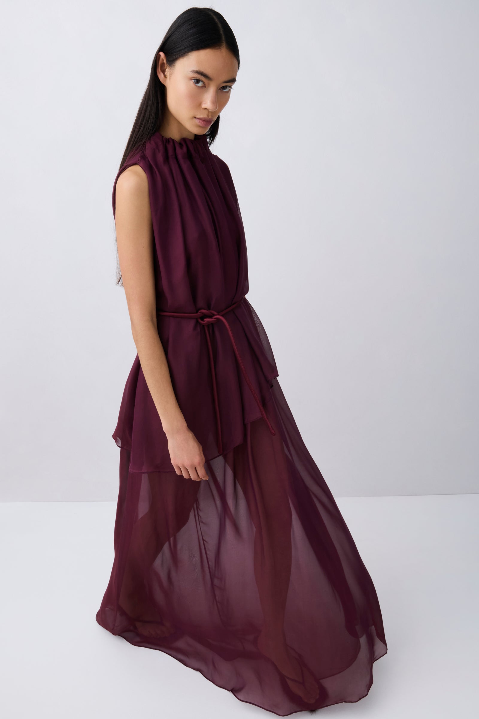 A woman modeling a sleeveless, high-neck, deep burgundy maxi dress with a matching waist tie, set against a plain light gray studio background.