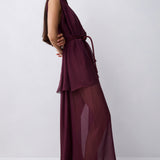 A woman wearing a sleeveless, high-neck, floor-length maxi dress in deep burgundy. The dress features a structured waist with a matching fabric tie belt and a flowing, semi-sheer chiffon skirt.