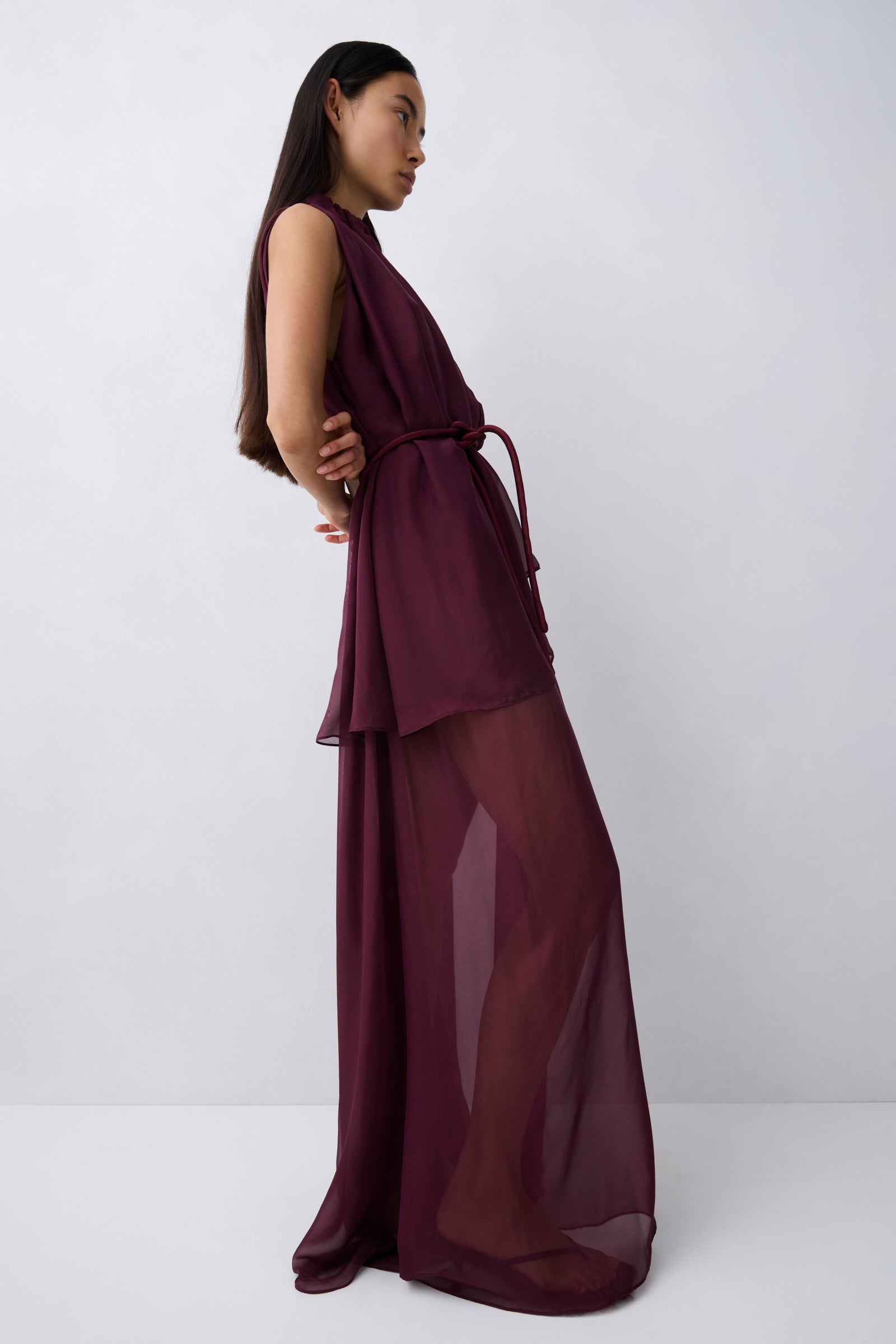 A woman wearing a sleeveless, high-neck, floor-length maxi dress in deep burgundy. The dress features a structured waist with a matching fabric tie belt and a flowing, semi-sheer chiffon skirt.