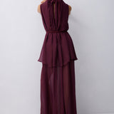 A woman viewed from behind wearing a sleeveless, deep burgundy maxi dress with a tie-neck back detail, a defined waist, and a flowing layered skirt, set against a plain light gray studio background.