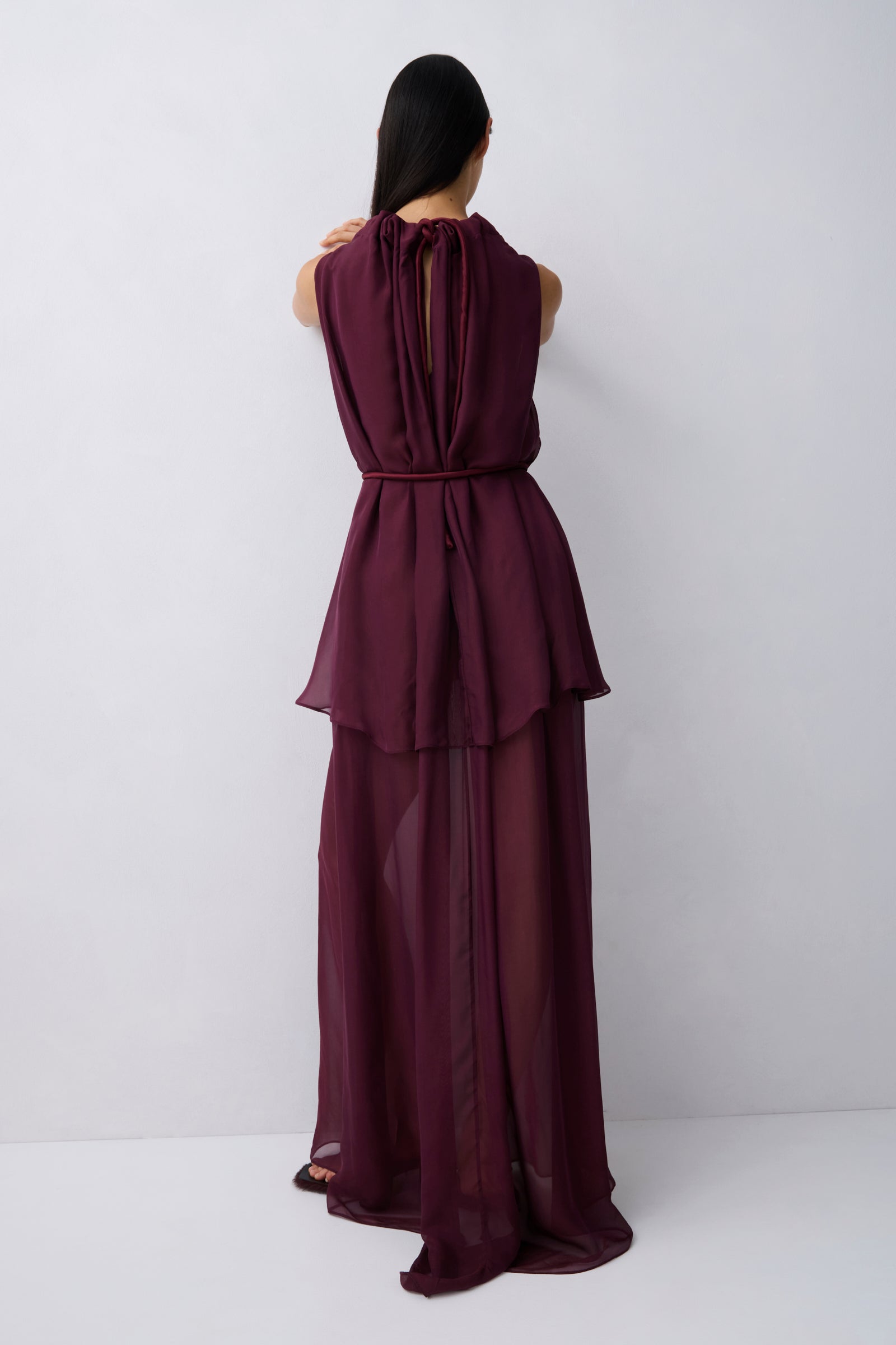 A woman viewed from behind wearing a sleeveless, deep burgundy maxi dress with a tie-neck back detail, a defined waist, and a flowing layered skirt, set against a plain light gray studio background.