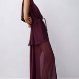 A model wearing a sleeveless, high-neck, deep burgundy maxi dress with a sheer, flowing overlay, photographed against a neutral light gray studio background.