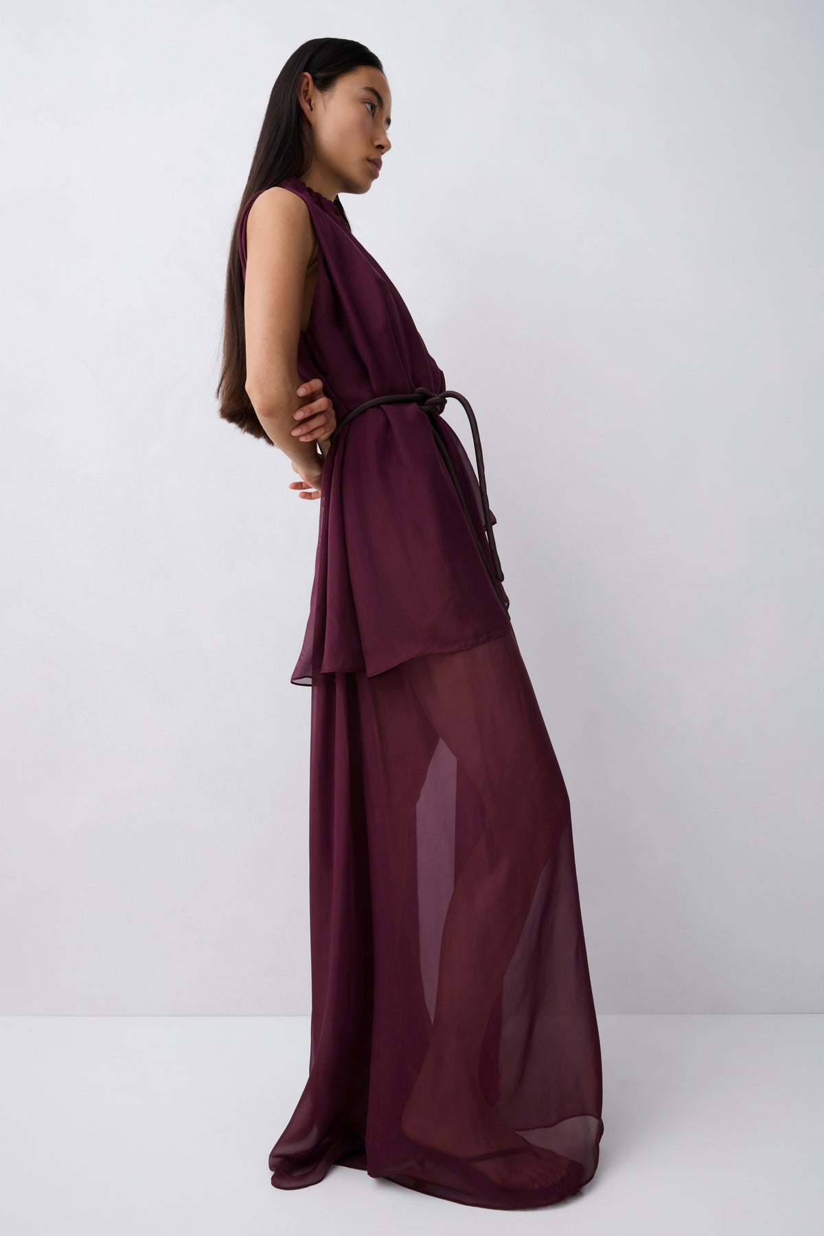 A model wearing a sleeveless, high-neck, deep burgundy maxi dress with a sheer, flowing overlay, photographed against a neutral light gray studio background.