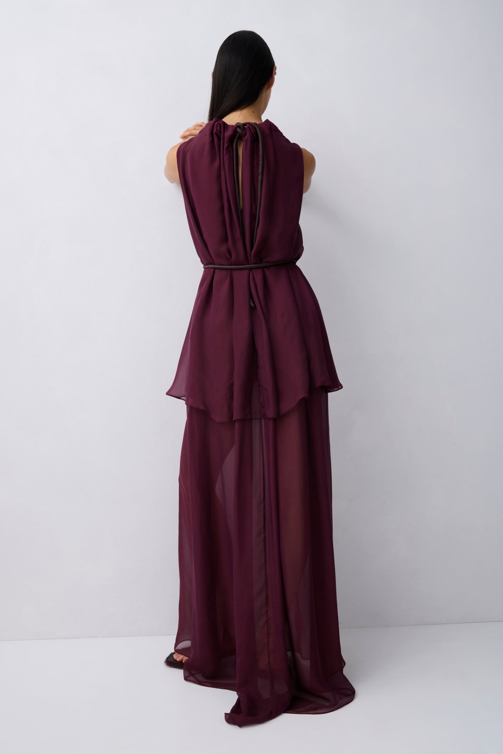 A model wearing a sleeveless, high-neck, deep burgundy maxi dress with a sheer, flowing overlay, photographed against a neutral light gray studio background.