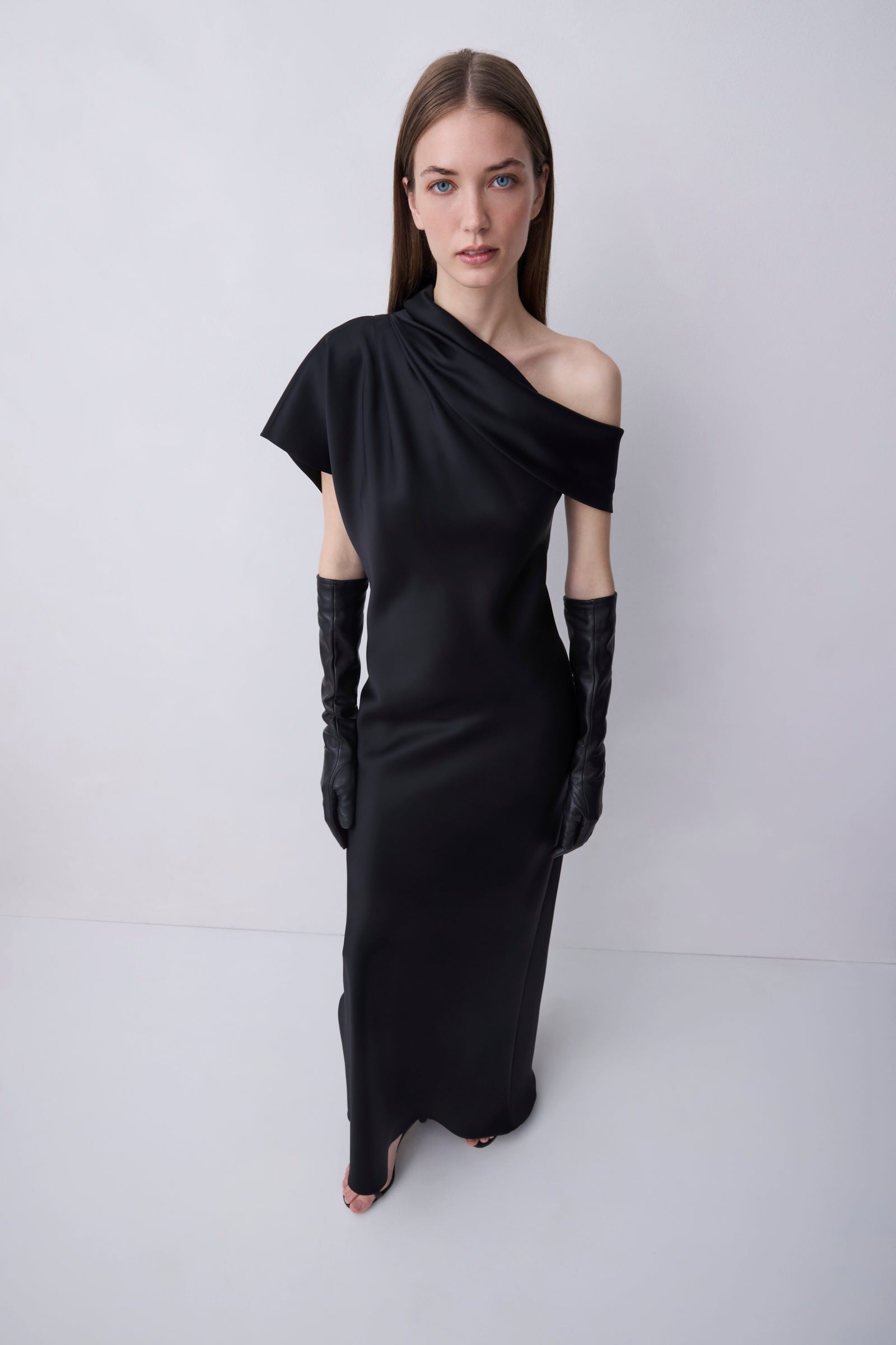 A model wearing a black one-shoulder satin dress with long black gloves, standing against a light gray studio background.