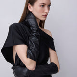 A model wearing a black one-shoulder dress with dramatic draped fabric and long black gloves, posed against a light gray studio background.