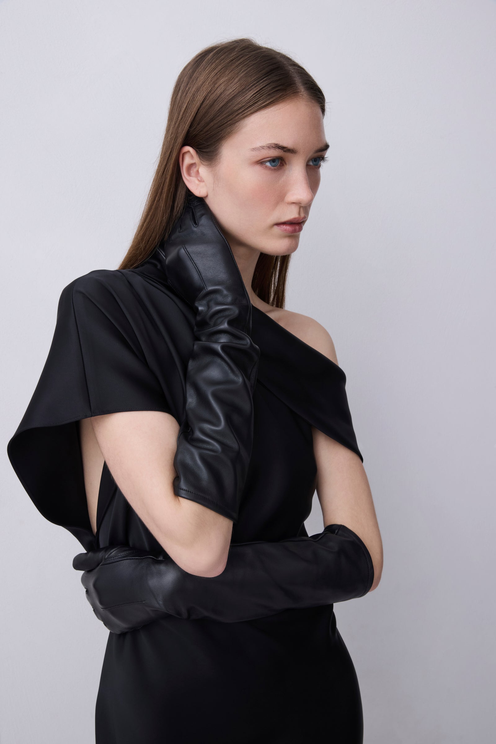 A model wearing a black one-shoulder dress with dramatic draped fabric and long black gloves, posed against a light gray studio background.