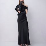 A woman modeling a black off-the-shoulder maxi dress with long black gloves and black strappy heels, standing against a light gray studio background.