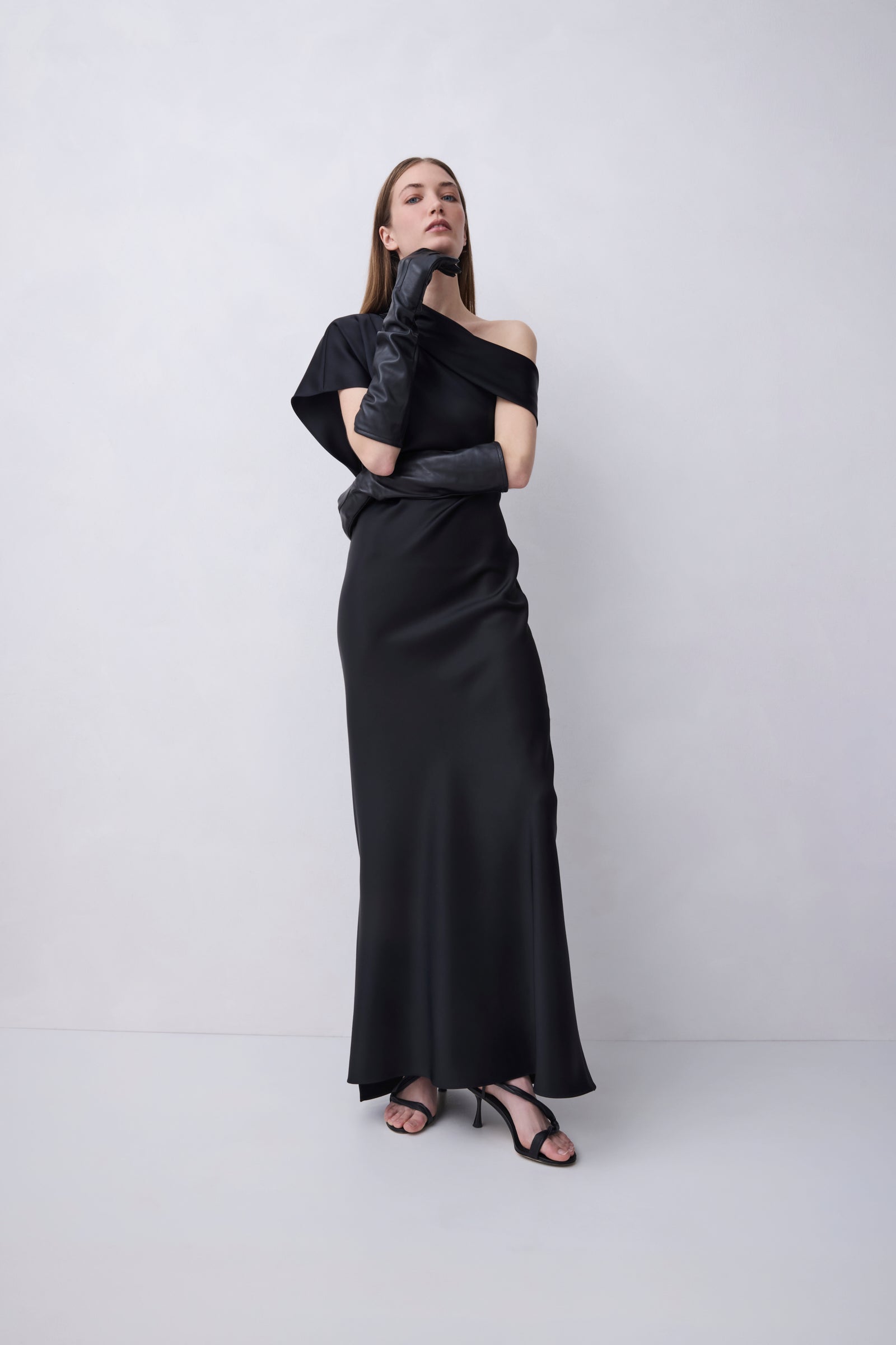 A woman modeling a black off-the-shoulder maxi dress with long black gloves and black strappy heels, standing against a light gray studio background.