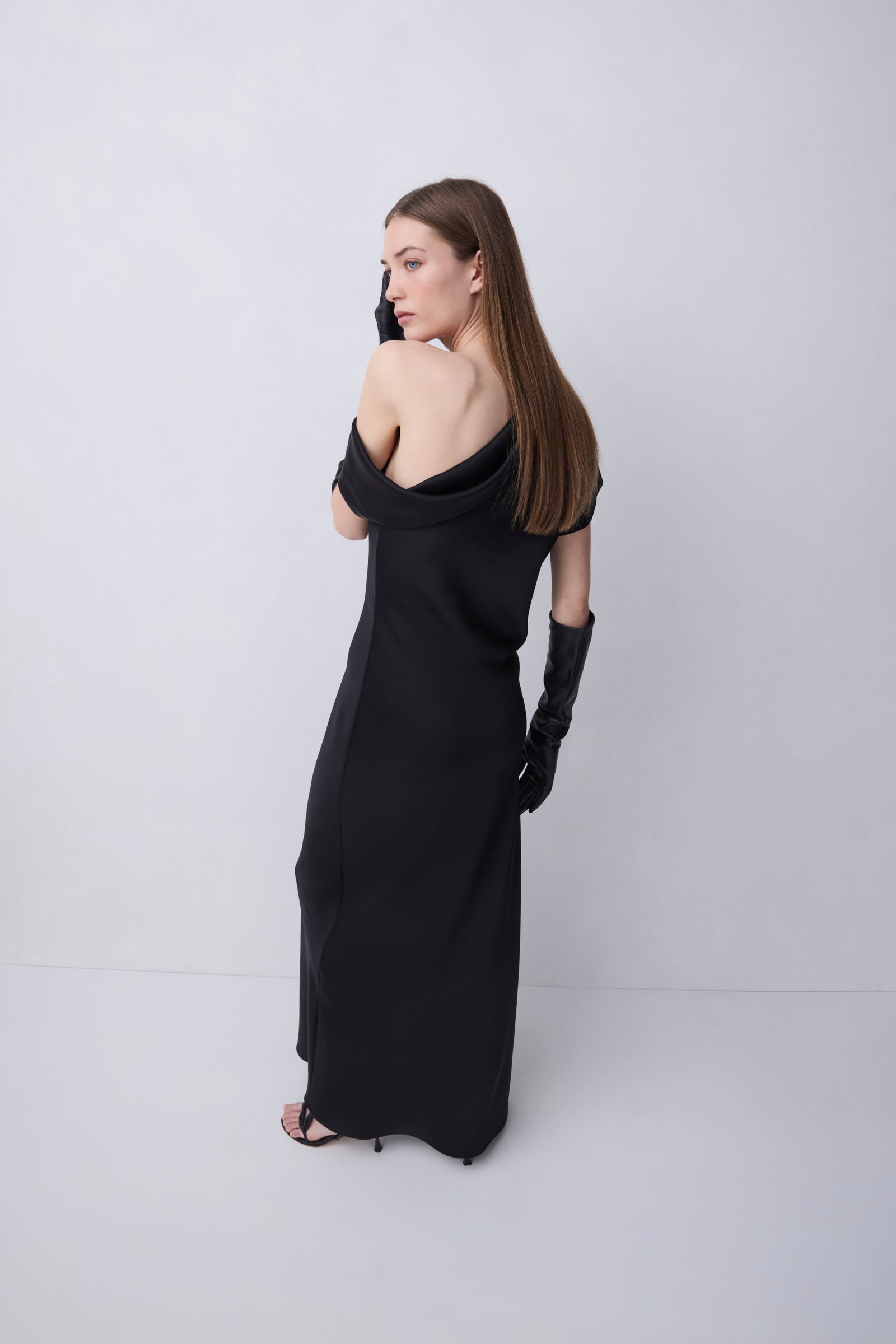 A woman modeling a black off-the-shoulder maxi dress with long black gloves, photographed from the back against a plain light gray studio background.