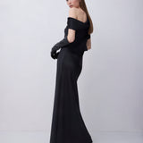 A woman modeling a floor-length black off-the-shoulder gown with long black gloves, photographed against a plain light gray studio background.