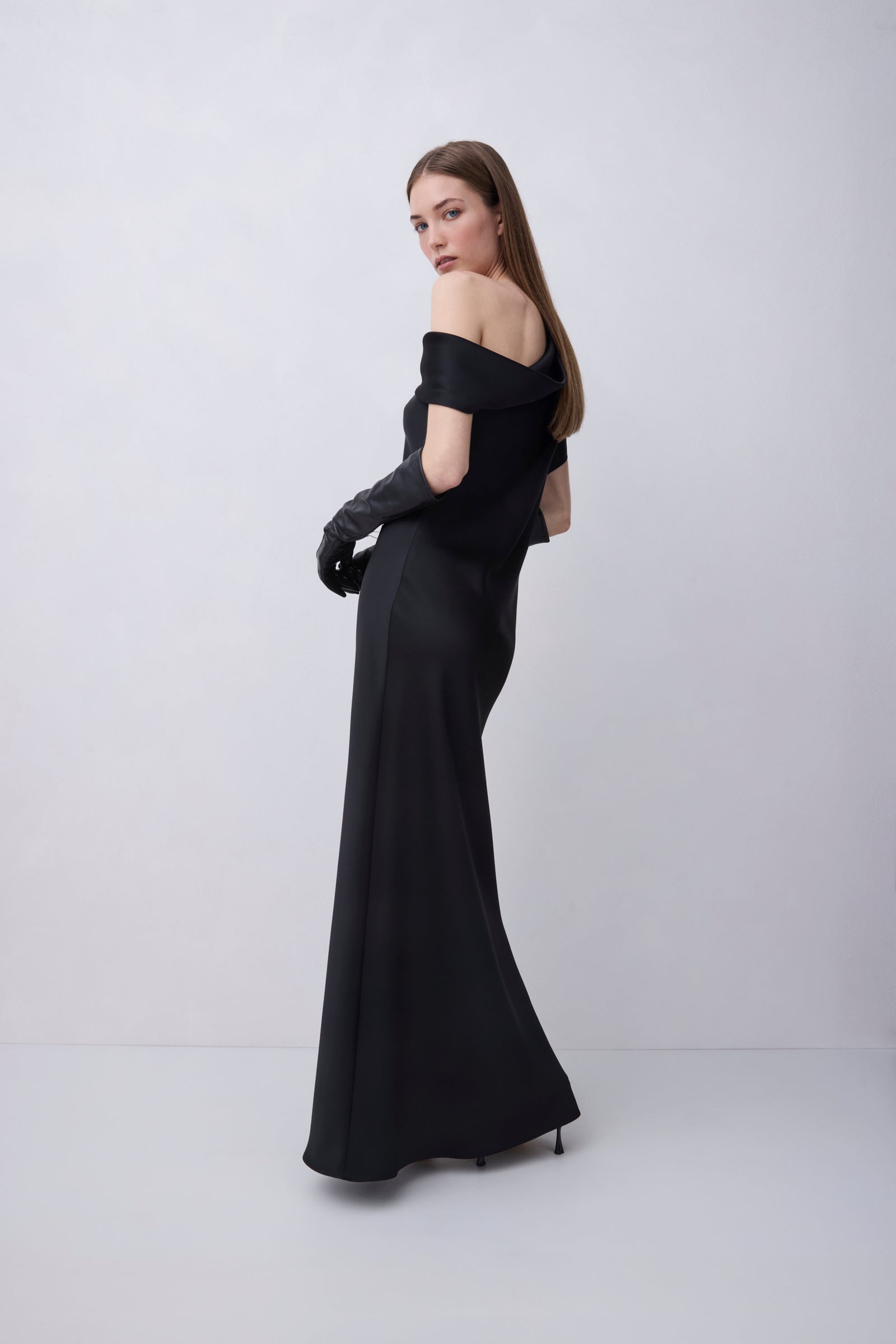 A woman modeling a floor-length black off-the-shoulder gown with long black gloves, photographed against a plain light gray studio background.