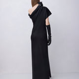 A model viewed from the back wearing a floor-length black one-shoulder gown with a draped neckline, short cape-like sleeves, and long black gloves, standing against a light gray studio background.