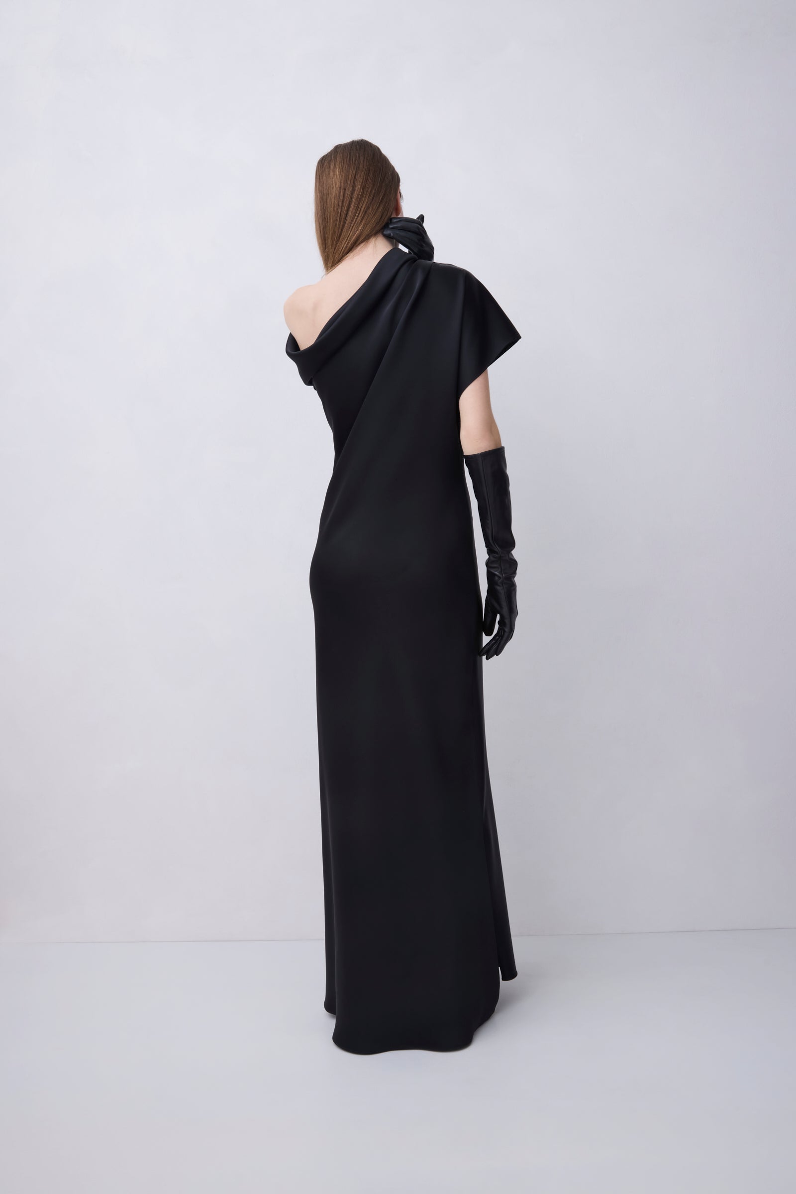 A model viewed from the back wearing a floor-length black one-shoulder gown with a draped neckline, short cape-like sleeves, and long black gloves, standing against a light gray studio background.