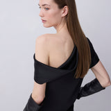A woman with long brown hair is shown from a side-back angle wearing a black off-the-shoulder dress with long black gloves, photographed against a plain light gray studio background.