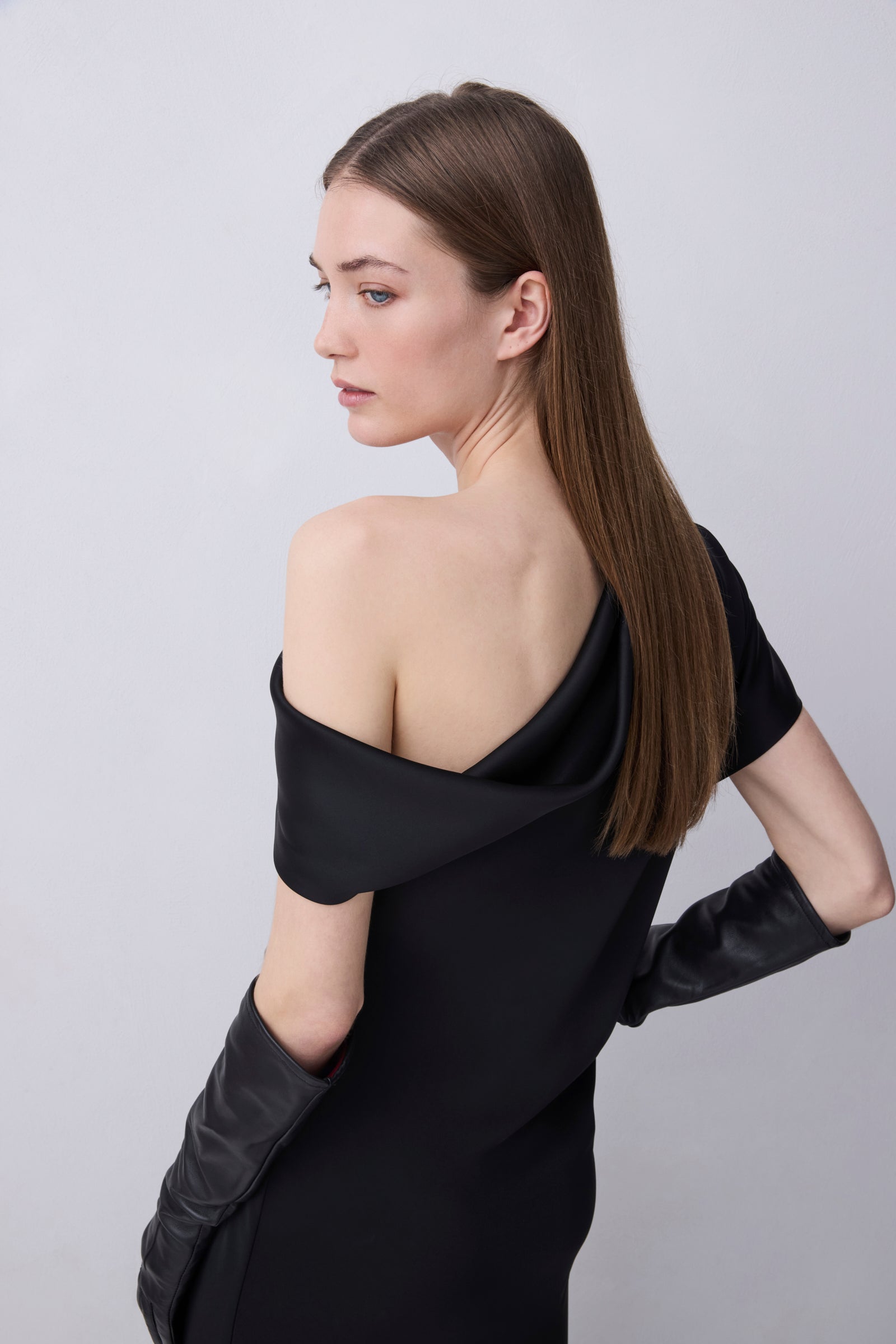 A woman with long brown hair is shown from a side-back angle wearing a black off-the-shoulder dress with long black gloves, photographed against a plain light gray studio background.