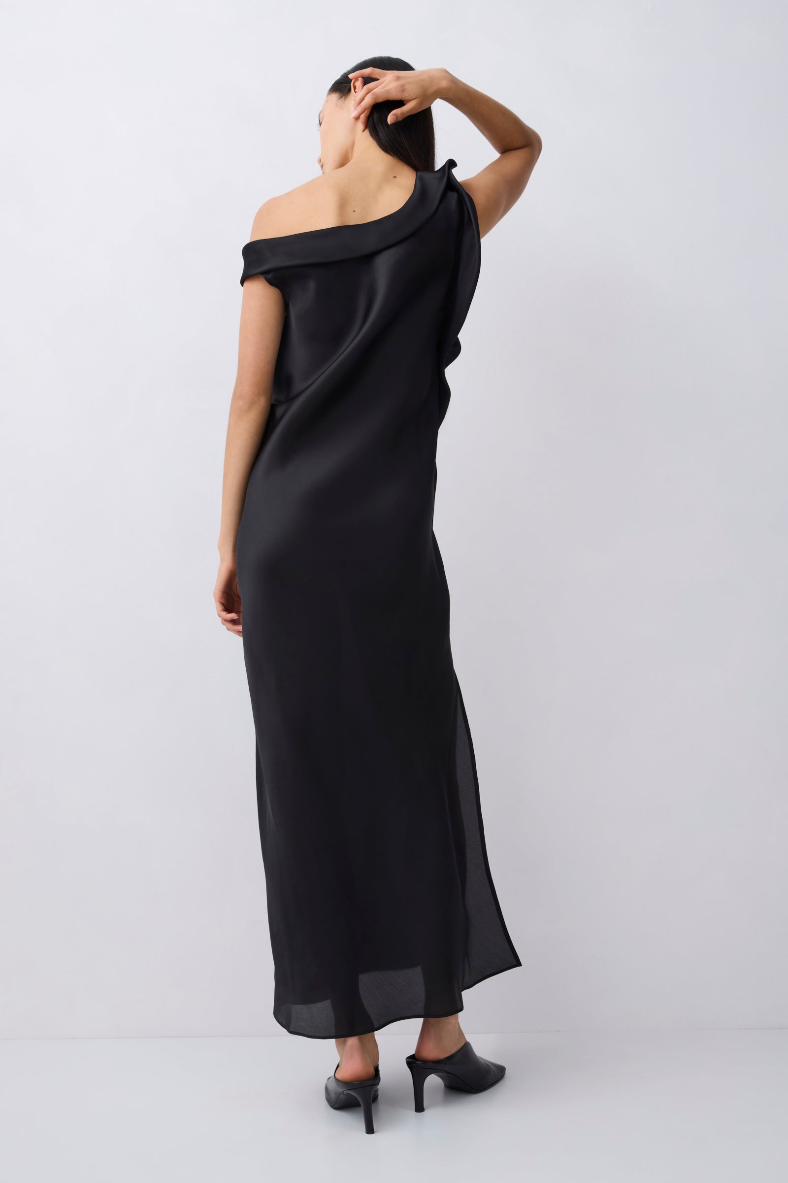 A woman wearing a black off-the-shoulder maxi dress with a side slit, shown from the back against a light gray studio background.