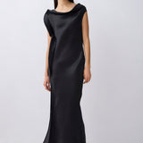 A model wearing a black satin slip dress with a draped, wide collar and cap sleeves, styled with black pointed-toe heels against a light gray studio background.