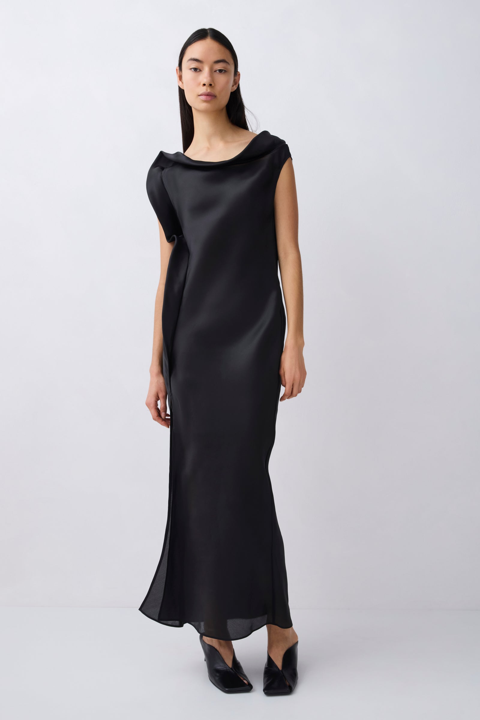 A model wearing a black satin slip dress with a draped, wide collar and cap sleeves, styled with black pointed-toe heels against a light gray studio background.