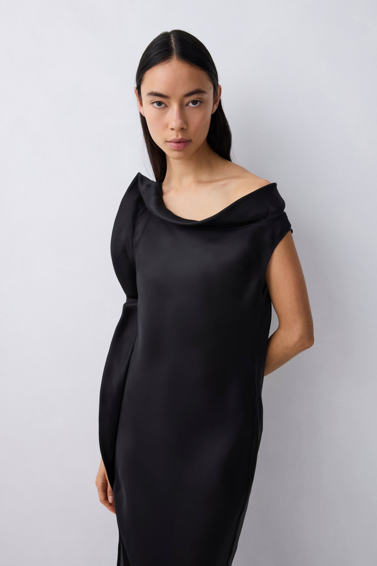 A woman modeling a black off-the-shoulder cowl-neck dress against a light gray studio background.