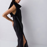 A model wearing a black sleeveless wrap-style maxi dress with a high slit and structured shoulders, paired with black pointed flats, set against a plain light gray studio background.