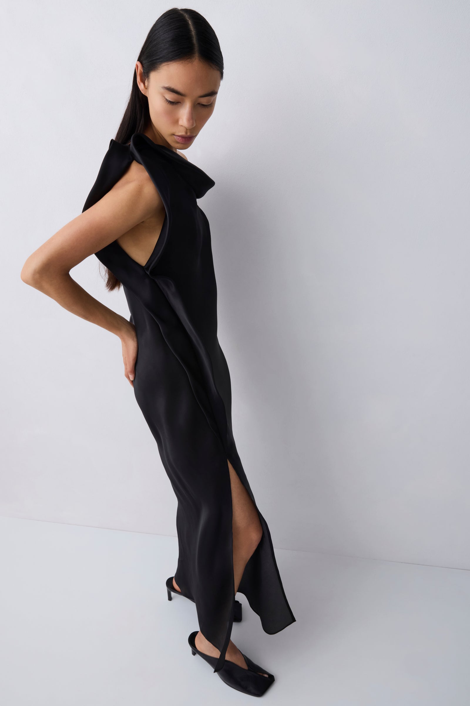 A model wearing a black sleeveless wrap-style maxi dress with a high slit and structured shoulders, paired with black pointed flats, set against a plain light gray studio background.