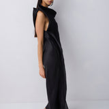 A model wearing a black sleeveless wrap-style maxi dress with a high draped collar and side slit, paired with black pointed-toe heels, standing against a light gray studio background.