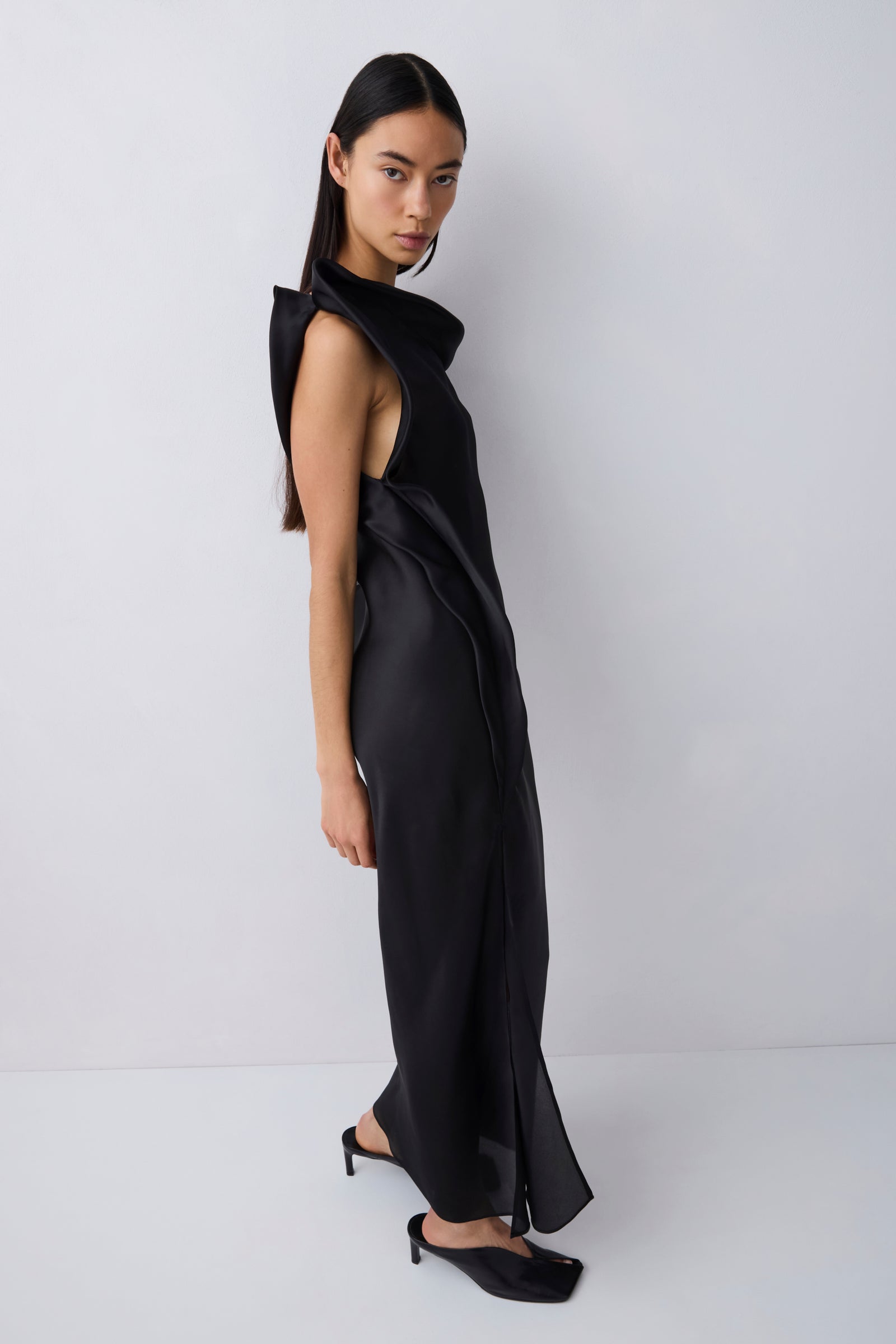 A model wearing a black sleeveless wrap-style maxi dress with a high draped collar and side slit, paired with black pointed-toe heels, standing against a light gray studio background.