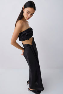 A model wearing a black strapless draped crop top and matching wide-leg draped trousers, styled with black pointed-toe heels against a light gray studio background.