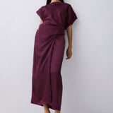 A model wearing a dark plum purple draped midi dress with short wide sleeves and black pointed-toe flats, standing against a light gray studio background.