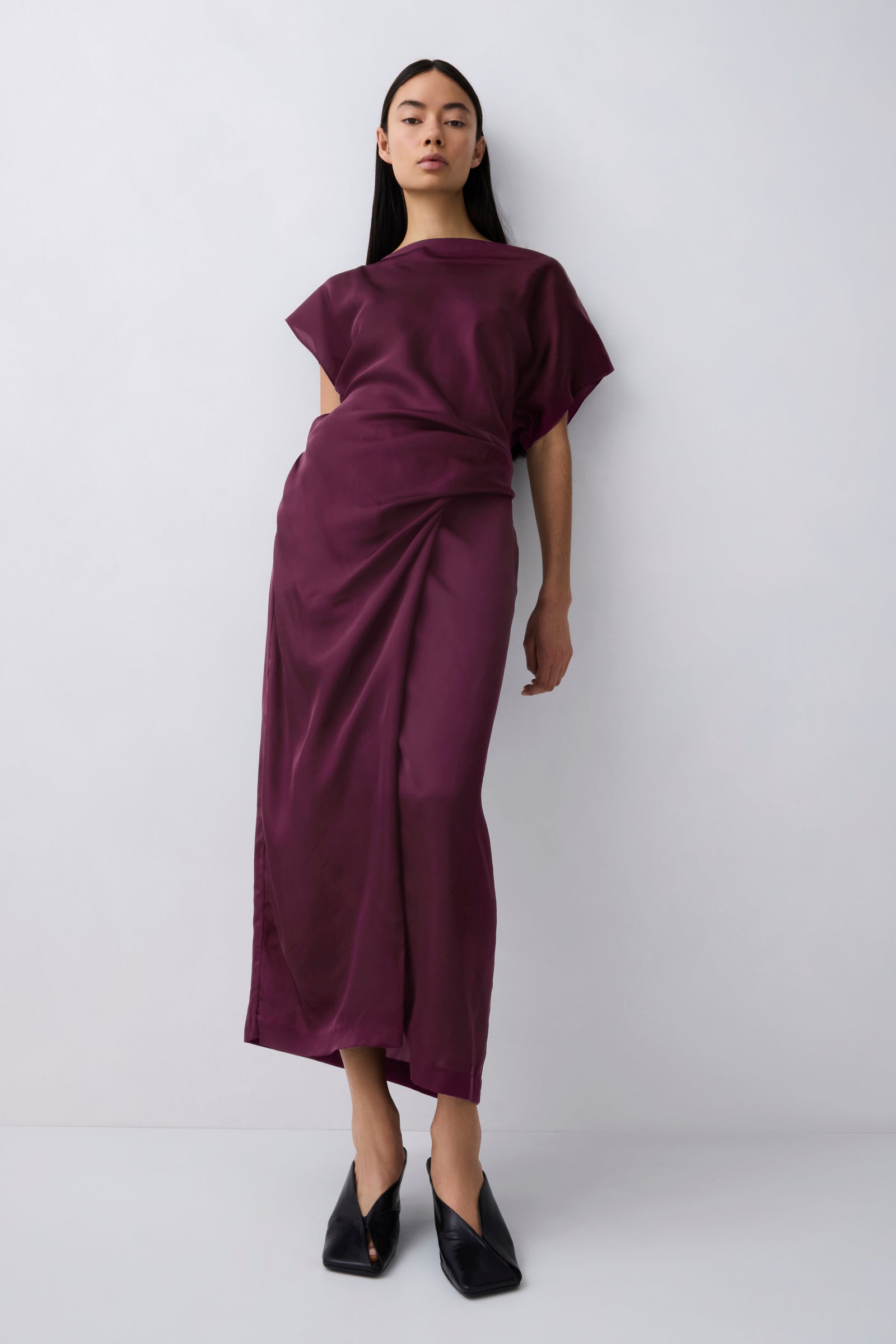 A model wearing a dark plum purple draped midi dress with short wide sleeves and black pointed-toe flats, standing against a light gray studio background.
