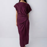 A woman modeling a draped, off-the-shoulder midi dress in deep burgundy, paired with black pointed-toe heels, against a plain light gray studio background.