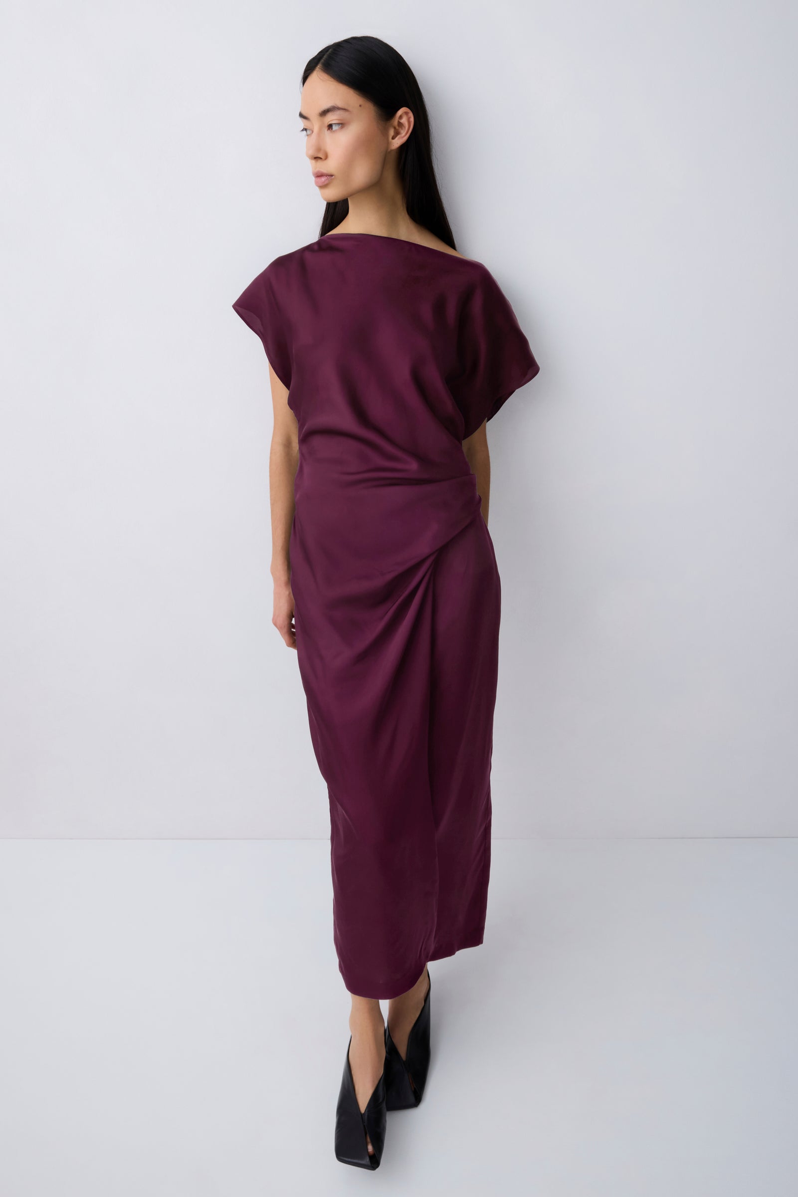 A woman modeling a draped, off-the-shoulder midi dress in deep burgundy, paired with black pointed-toe heels, against a plain light gray studio background.