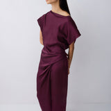 A woman modeling a draped, off-the-shoulder maxi dress in deep burgundy with a gathered waist, paired with black pointed flats, against a light gray studio background.