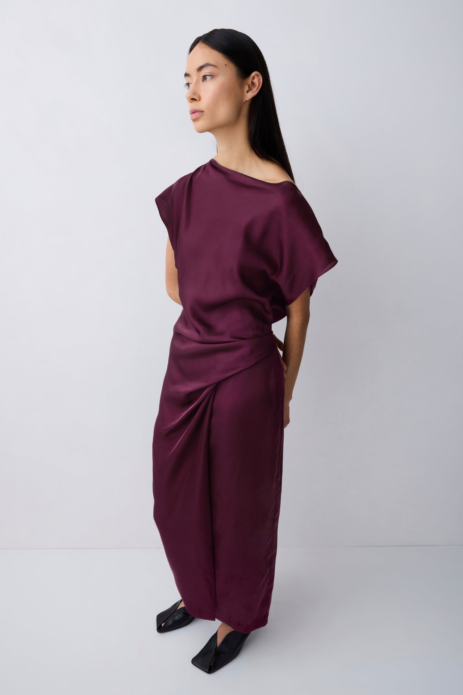 A woman modeling a draped, off-the-shoulder maxi dress in deep burgundy with a gathered waist, paired with black pointed flats, against a light gray studio background.