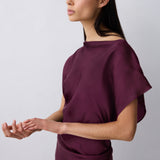 A woman modeling a draped, off-the-shoulder wrap-style top in deep plum purple against a light gray studio background.