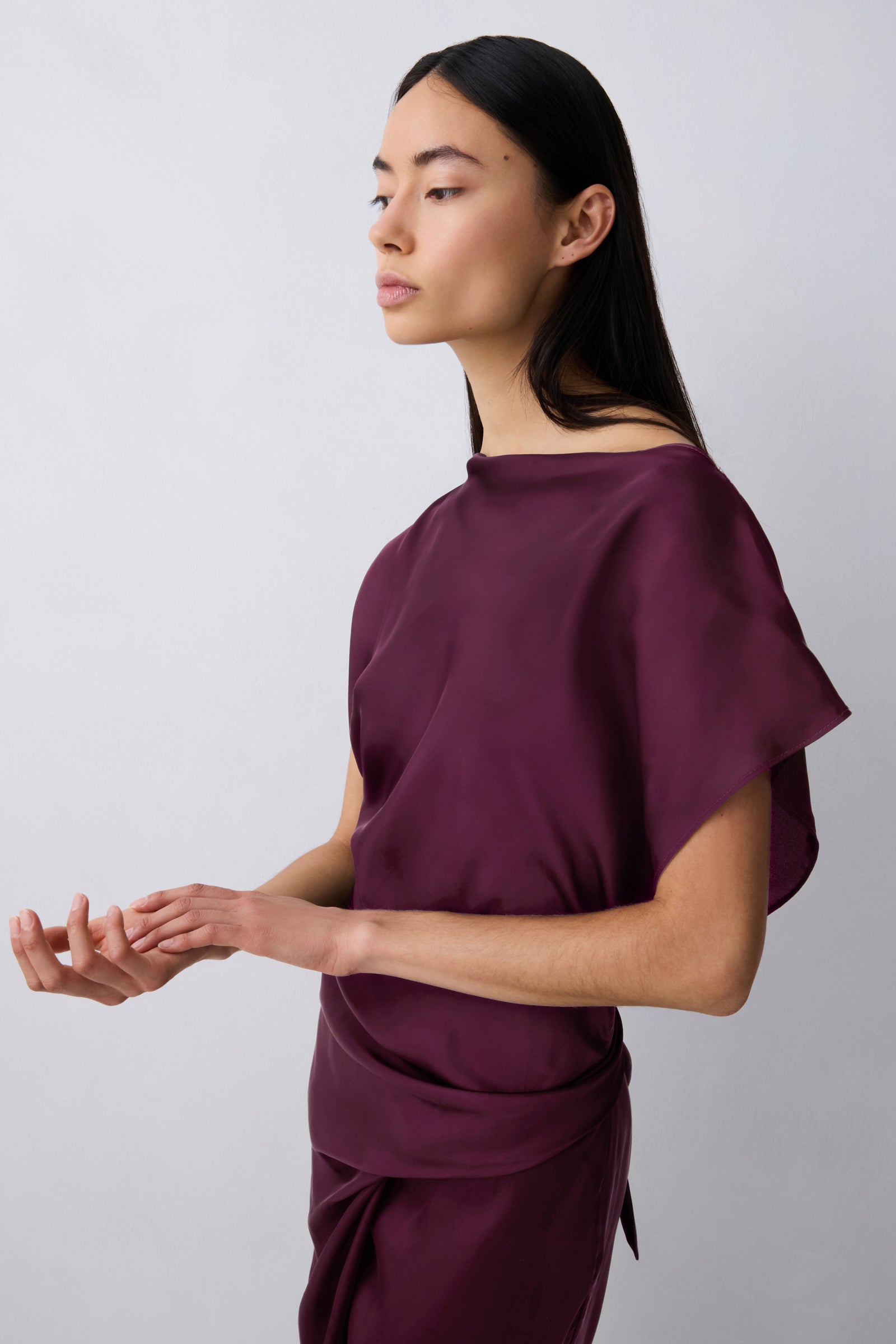 A woman modeling a draped, off-the-shoulder wrap-style top in deep plum purple against a light gray studio background.