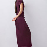 A woman wearing a sleeveless, deep plum-colored midi dress with puff sleeves and a side tie, paired with black mule heels, shown from a side profile against a light gray studio background.