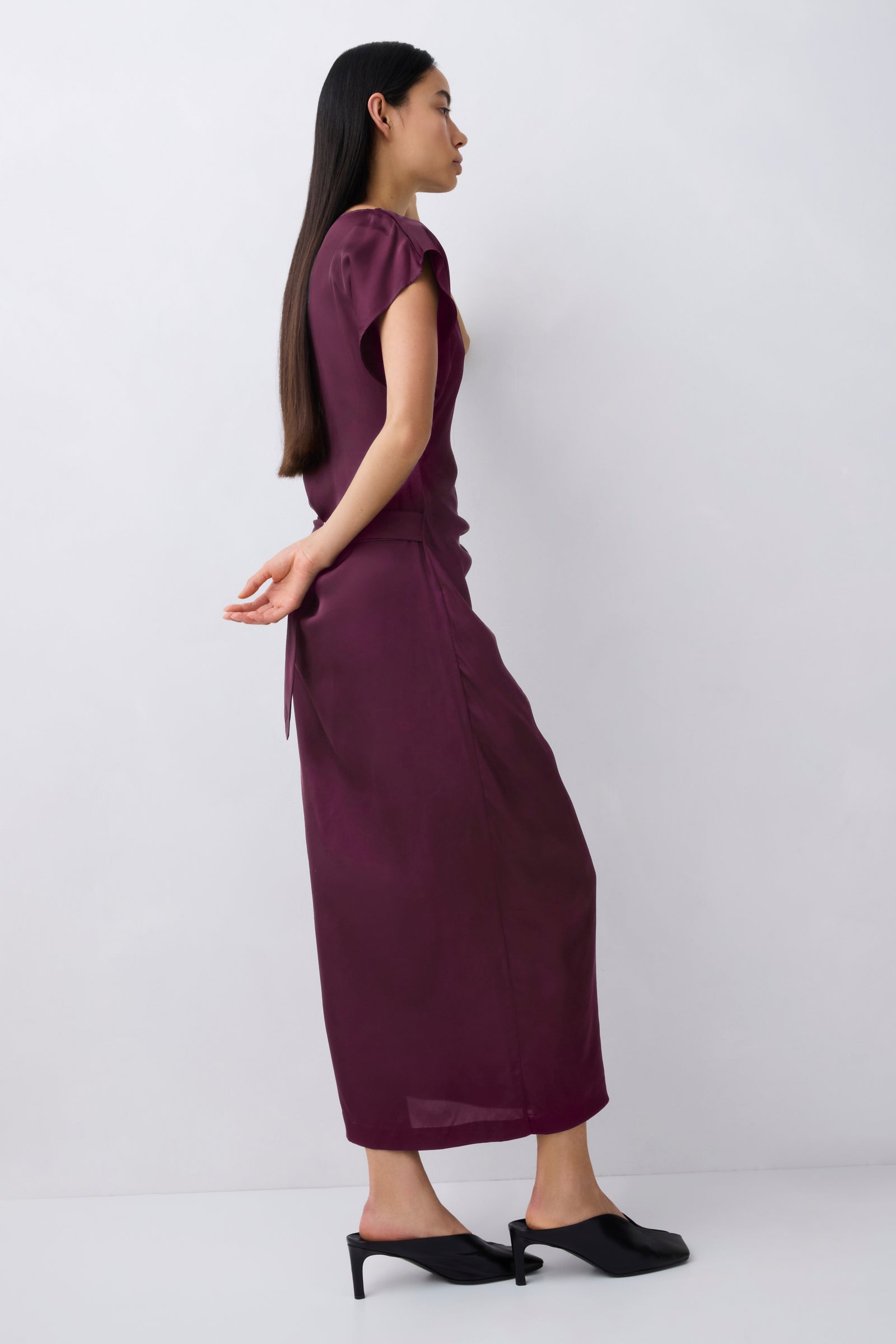 A woman wearing a sleeveless, deep plum-colored midi dress with puff sleeves and a side tie, paired with black mule heels, shown from a side profile against a light gray studio background.