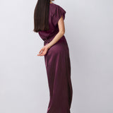 A woman with long dark hair is shown from the back wearing a satin-like dark plum midi dress with short puff sleeves and black mule heels, standing against a light gray studio background.