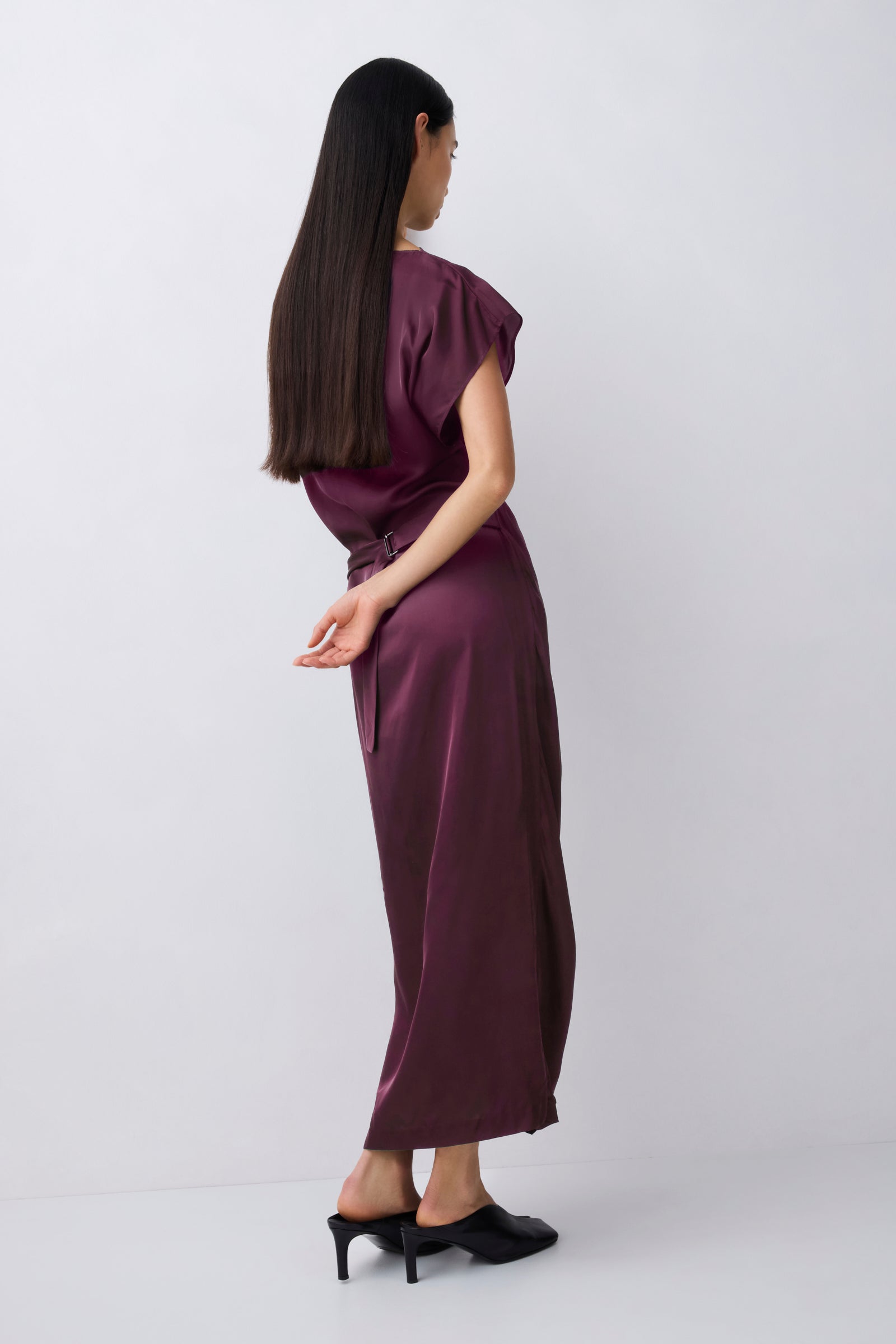 A woman with long dark hair is shown from the back wearing a satin-like dark plum midi dress with short puff sleeves and black mule heels, standing against a light gray studio background.