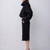A woman stands in profile wearing a black cropped jacket with a high structured collar and a matching black midi skirt, paired with black strappy heels, against a plain light gray studio background.