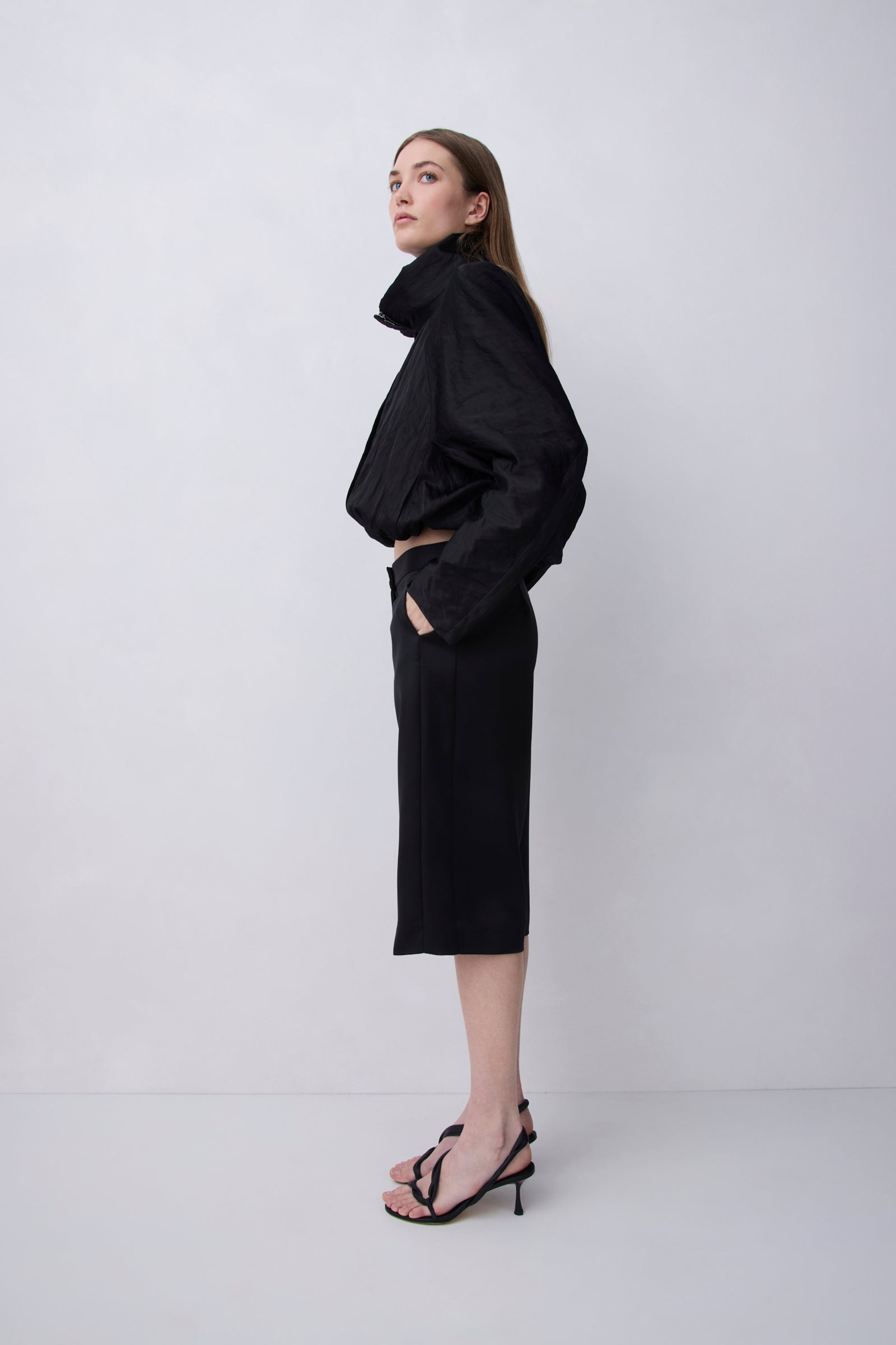 A woman stands in profile wearing a black cropped jacket with a high structured collar and a matching black midi skirt, paired with black strappy heels, against a plain light gray studio background.