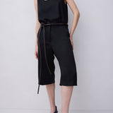 A model wearing a black sleeveless top and matching black cropped pants with a thin dark tie belt, paired with black strappy high-heeled sandals, against a light gray studio background.