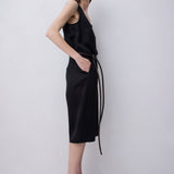 A woman modeling a black sleeveless midi dress with a thin waist tie, paired with black strappy high-heeled sandals, against a plain light gray studio background.