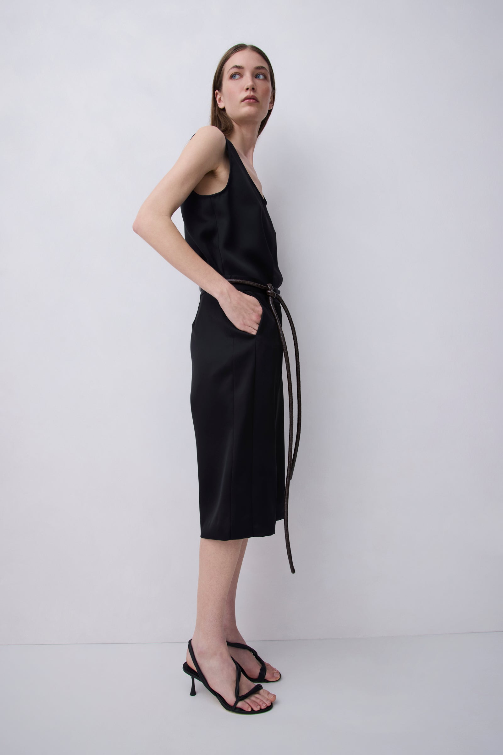 A woman modeling a black sleeveless midi dress with a thin waist tie, paired with black strappy high-heeled sandals, against a plain light gray studio background.